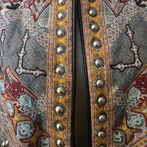 May include: Close-up of a gray jacket with intricate embroidered patterns in shades of red, gold, and teal. The jacket's edges are trimmed with silver metal studs and black leather. The overall design is ornate and detailed.
