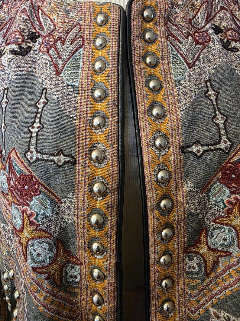 May include: Close-up of a gray jacket with intricate embroidered patterns in shades of red, gold, and teal. The jacket's edges are trimmed with silver metal studs and black leather. The overall design is ornate and detailed.