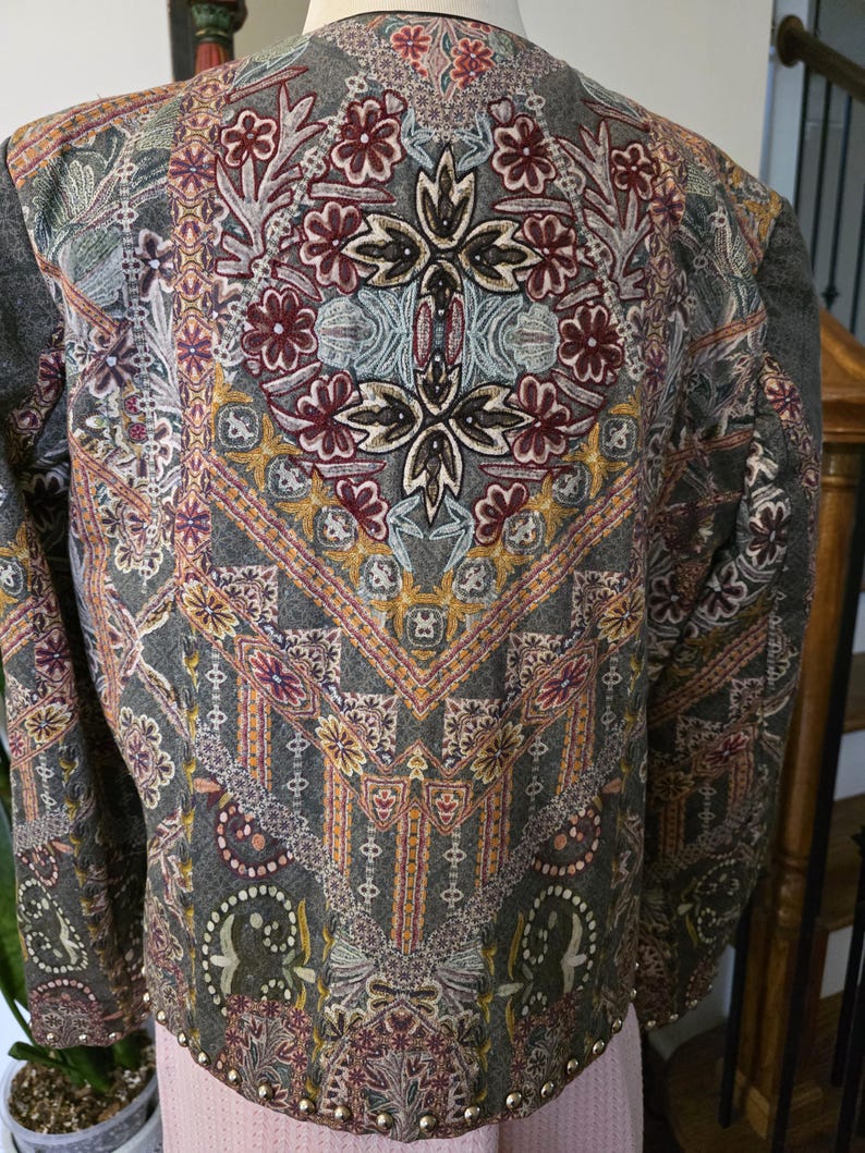 May include: A gray jacket with an intricate floral and geometric pattern in burgundy, gold, and teal. The jacket has a round neckline and is embellished with small, gold-colored studs along the bottom hem.