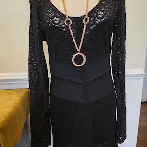May include: Black lace dress with long sleeves and a scoop neckline. A gold-colored necklace with multiple circular pendants hangs over the dress. The dress has a fitted waist and a textured pattern.