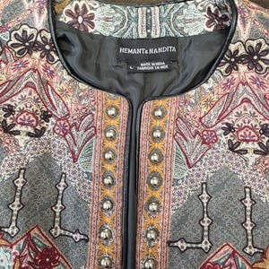 May include: Close-up of a decorative jacket with an intricate paisley pattern in shades of gray, burgundy, and gold. The jacket features a black trim and silver button closures. The label reads "HEMANT & NANDITA".