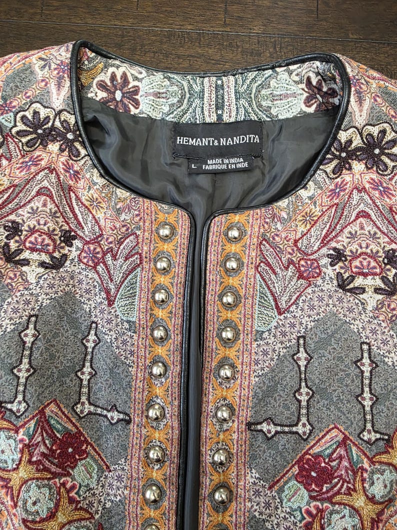 May include: Close-up of a decorative jacket with an intricate paisley pattern in shades of gray, burgundy, and gold. The jacket features a black trim and silver button closures. The label reads "HEMANT & NANDITA".