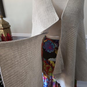 May include: A light grey, ribbed knit cardigan with a draped, open front. The cardigan is displayed on a mannequin, revealing a colourful floral skirt underneath. A gold-toned necklace is visible at the neckline.