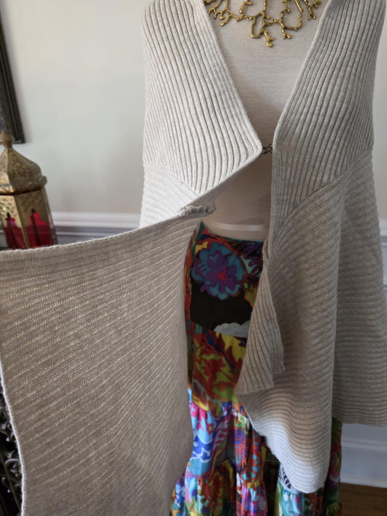 May include: A light grey, ribbed knit cardigan with a draped, open front. The cardigan is displayed on a mannequin, revealing a colourful floral skirt underneath. A gold-toned necklace is visible at the neckline.