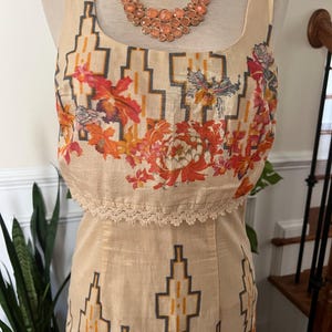 May include: A beige sleeveless dress with a geometric pattern and floral accents. The top portion features a square neckline and a lace trim. A peach-colored necklace with large stones is displayed on a mannequin.