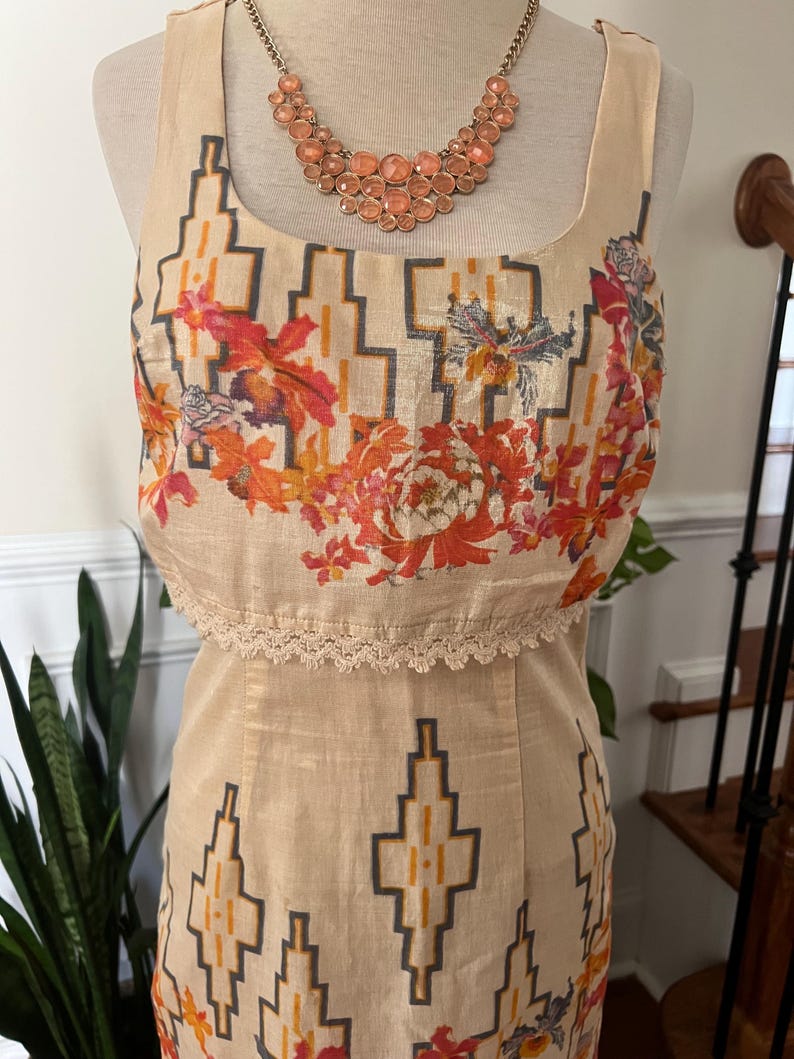 May include: A beige sleeveless dress with a geometric pattern and floral accents. The top portion features a square neckline and a lace trim. A peach-colored necklace with large stones is displayed on a mannequin.