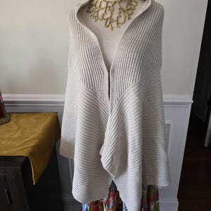 May include: A beige ribbed knit hooded cape with a draped front, displayed on a mannequin. A gold-coloured statement necklace is worn underneath the hood. The cape is paired with a colourful patterned skirt.
