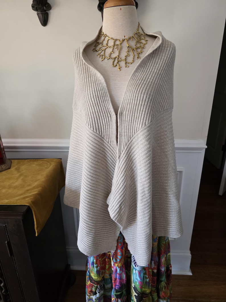 May include: A beige ribbed knit hooded cape with a draped front, displayed on a mannequin. A gold-coloured statement necklace is worn underneath the hood. The cape is paired with a colourful patterned skirt.