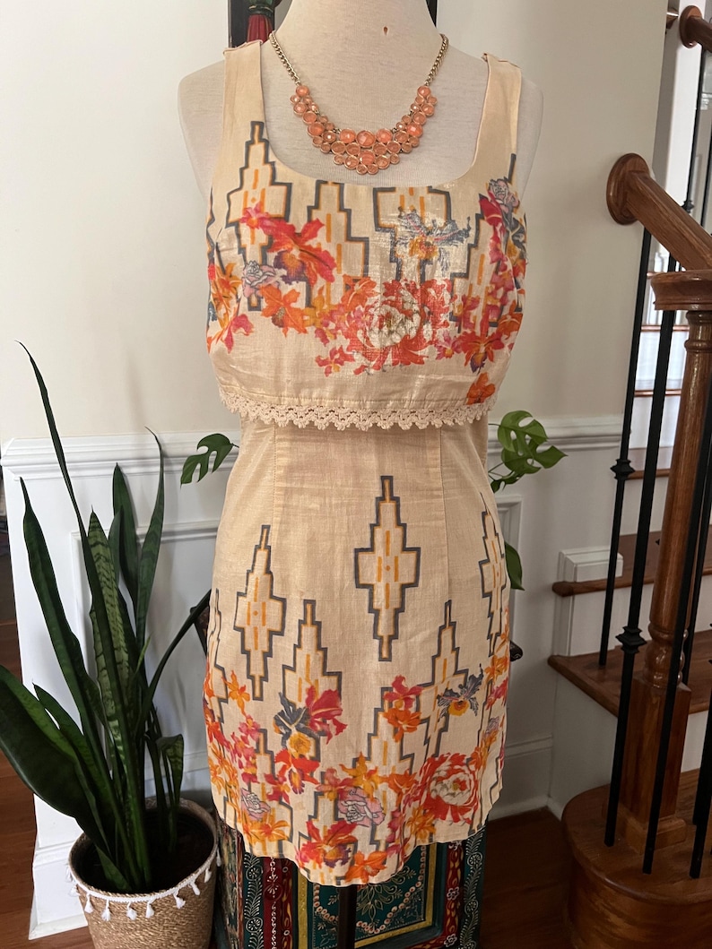 May include: A two-piece dress on a mannequin. The top is a square-neck tank with a floral print in orange, red, and purple. The skirt has a geometric pattern in beige and brown. A peach-colored beaded necklace is around the mannequin's neck.