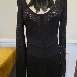 May include: Black lace dress with long sleeves and a scoop neckline. The dress features a fitted bodice and a flared skirt. A black feather necklace is draped around the neckline. The dress is displayed on a mannequin.