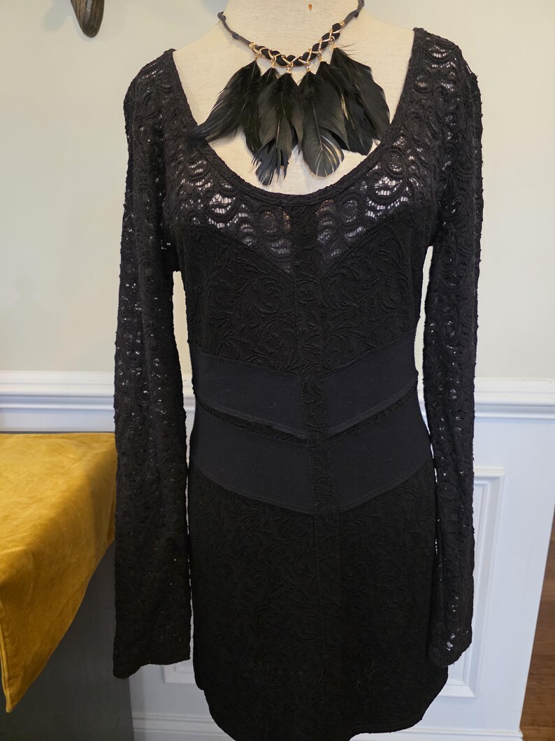 May include: Black lace dress with long sleeves and a scoop neckline. The dress features a fitted bodice and a flared skirt. A black feather necklace is draped around the neckline. The dress is displayed on a mannequin.