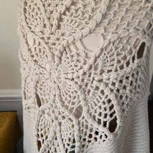 May include: Cream-coloured, open-weave crochet vest with a detailed, fan-shaped design on the back. The vest has a ribbed knit bottom and a sleeveless design. The crochet pattern features intricate openwork and textured stitches, creating a unique, handmade look.