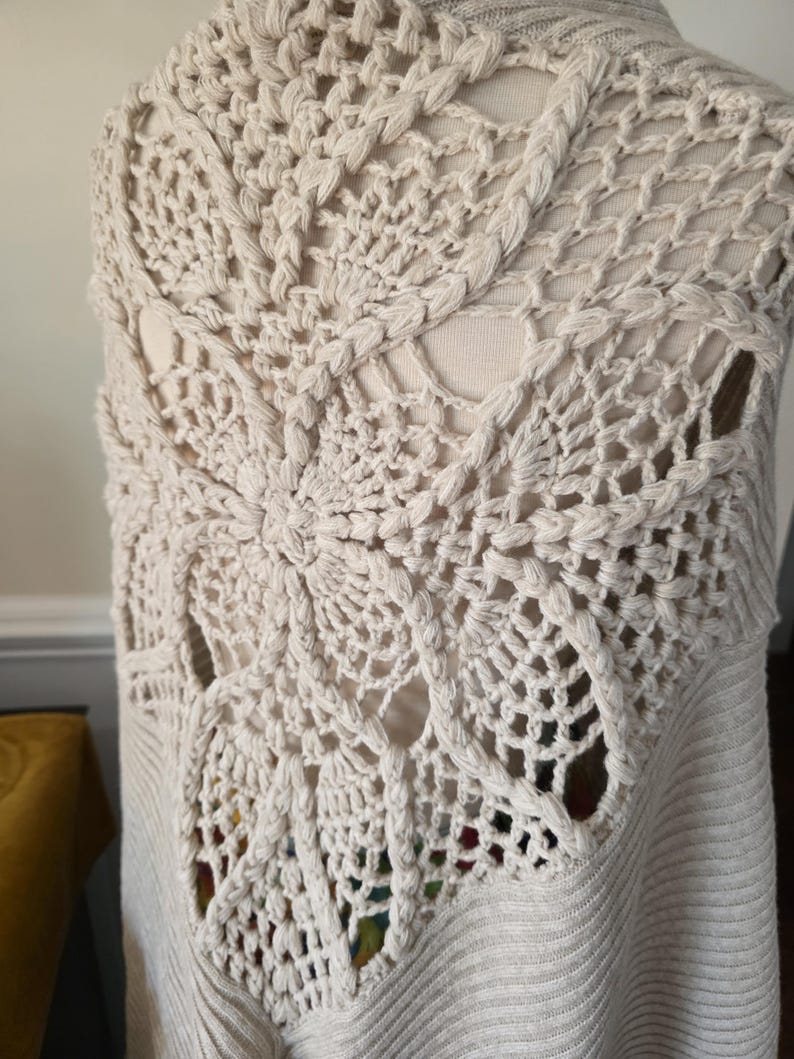 May include: Cream-coloured, open-weave crochet vest with a detailed, fan-shaped design on the back. The vest has a ribbed knit bottom and a sleeveless design. The crochet pattern features intricate openwork and textured stitches, creating a unique, handmade look.