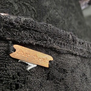 May include: Close-up of a black garment with a textured pattern. A brown tag with the words "Free People" is attached to the garment. The fabric appears to be a knit or crochet material.