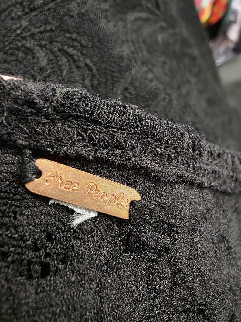 May include: Close-up of a black garment with a textured pattern. A brown tag with the words "Free People" is attached to the garment. The fabric appears to be a knit or crochet material.