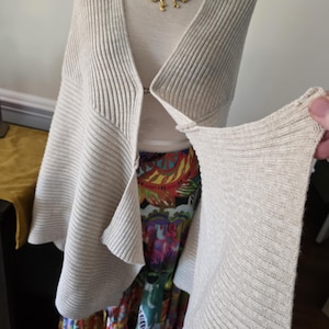May include: A light beige, ribbed knit cardigan with a draped front is displayed on a mannequin. The cardigan is paired with a colourful patterned skirt and a gold-toned statement necklace. The sleeve of the cardigan is being held up.
