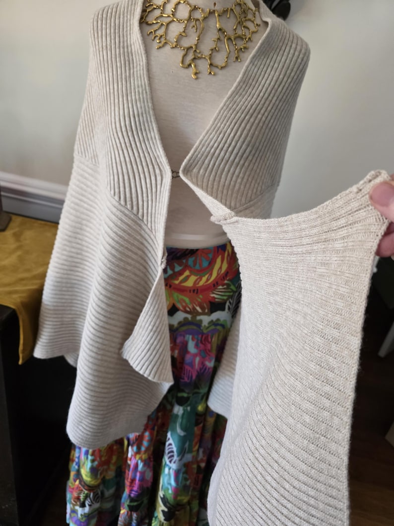 May include: A light beige, ribbed knit cardigan with a draped front is displayed on a mannequin. The cardigan is paired with a colourful patterned skirt and a gold-toned statement necklace. The sleeve of the cardigan is being held up.