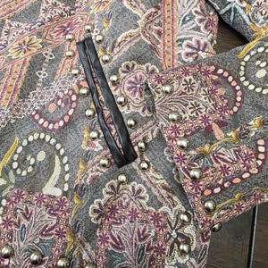 May include: Close-up of a gray jacket with a paisley pattern in shades of purple, orange, and gold. The jacket features silver stud embellishments along the edges and a black leather trim. The fabric has a textured appearance.