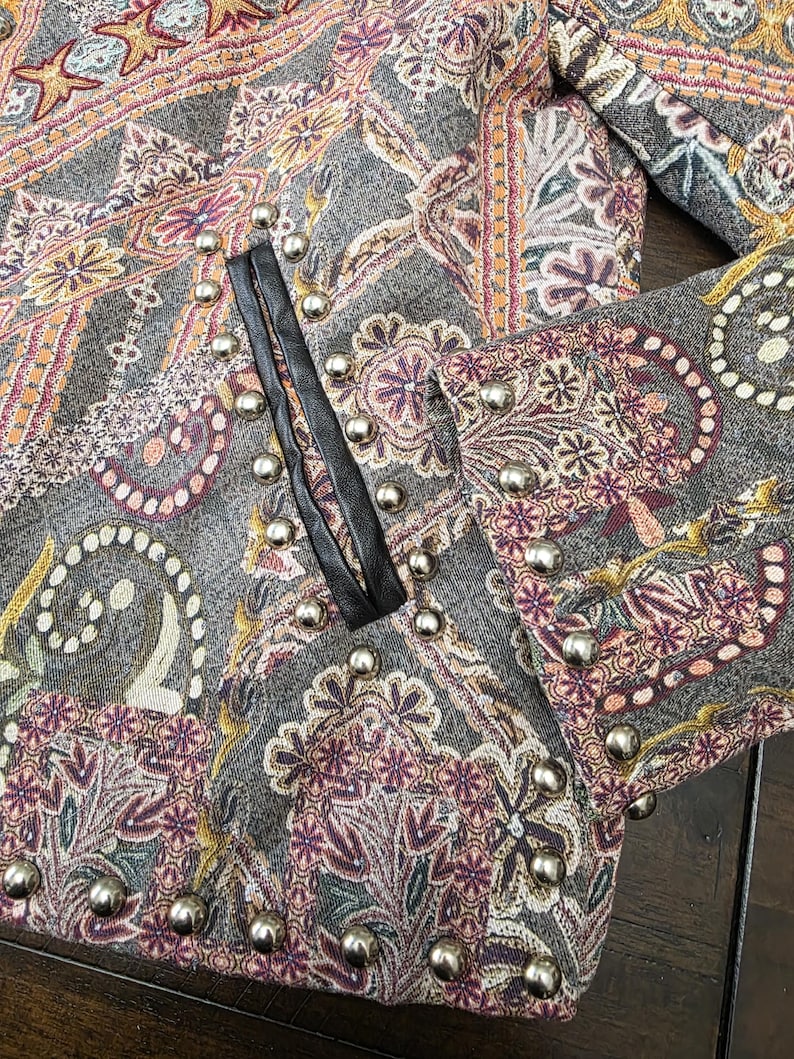 May include: Close-up of a gray jacket with a paisley pattern in shades of purple, orange, and gold. The jacket features silver stud embellishments along the edges and a black leather trim. The fabric has a textured appearance.