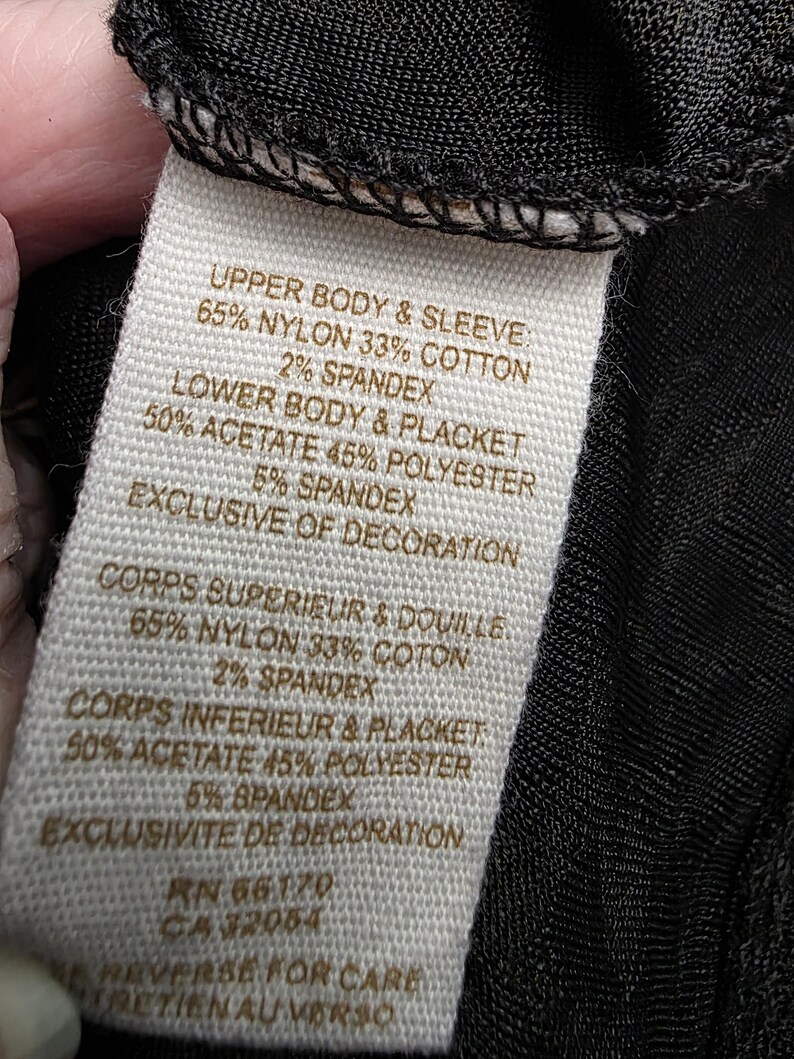 May include: A close-up of a clothing tag with text detailing the fabric composition. The tag lists the materials as nylon, cotton, acetate, polyester, and spandex. The tag also includes French translations of the fabric details.