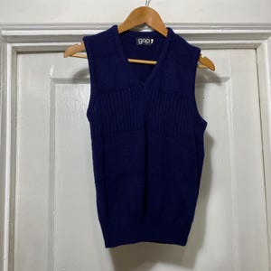 May include: A navy blue Gap brand V-neck sweater vest. The vest is hanging on a wooden hanger. The vest has a ribbed texture and a V-neck design. The Gap logo is visible on the upper back of the vest.