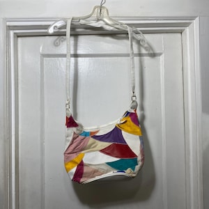 Könnte beinhalten: Eine weiße Crossbody-Tasche aus Leder mit Patchwork-Design. Die Tasche hat eine Vielzahl von farbenfrohen Lederstücken in Rosa, Gelb, Lila, Rot und Türkis. Die Tasche hat einen weißen Riemen und hängt an einem weißen Kleiderbügel.