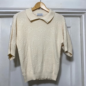 Pode incluir: Um suéter de cor creme de manga curta com gola e textura de malha waffle. O suéter tem um padrão geométrico e uma etiqueta que diz "Gabbie Kule".