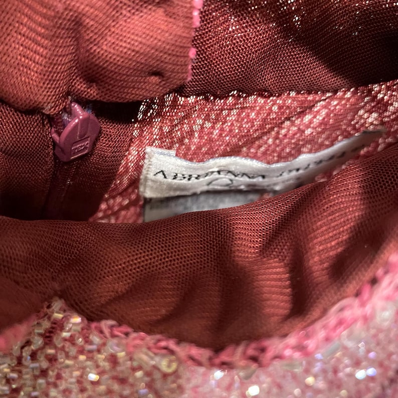 May include: Close-up of a burgundy dress with a zipper and a tag that reads "ABRIANNA". The dress has a mesh overlay and is embellished with pink and white beads. The zipper is a matching burgundy color.