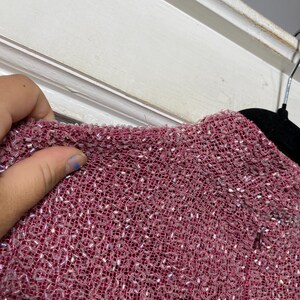 May include: Close-up of a textured, reddish-pink fabric, densely covered with small, shiny, silver-colored beads. The fabric appears to be woven, with a slightly rough texture. The image shows a portion of a garment.