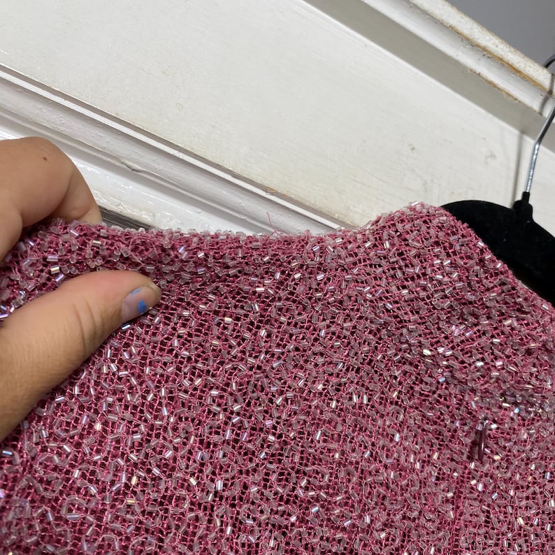 May include: Close-up of a textured, reddish-pink fabric, densely covered with small, shiny, silver-colored beads. The fabric appears to be woven, with a slightly rough texture. The image shows a portion of a garment.
