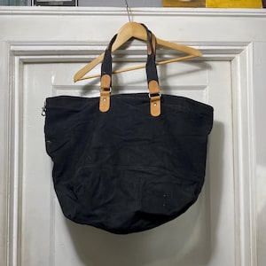 May include: A black tote bag with brown leather handles and accents, hanging on a wooden hanger. The bag has a zippered top and a large, open interior. The handles are attached with brass-colored hardware.