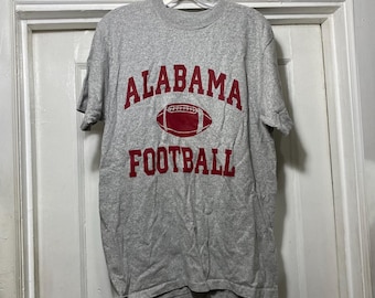 May include: A gray t-shirt with the words "ALABAMA FOOTBALL" in red block letters and a red football graphic. The shirt is hanging on a plastic hanger.