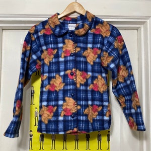 Peut inclure: Une veste en polaire bleu et blanc à carreaux avec un imprimé d'ours en peluche brun. La veste a une fermeture boutonnée et un col. L'étiquette indique "Disney".
