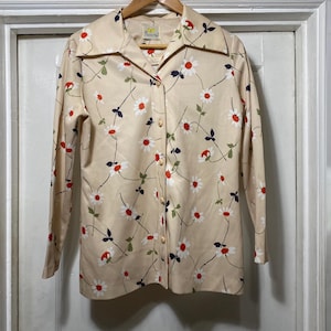 May include: A vintage button-down shirt in a cream color, adorned with a floral pattern of white daisies with red centers and green leaves. The long-sleeved shirt has a collared neckline and is buttoned up. The shirt is hanging on a wooden hanger.