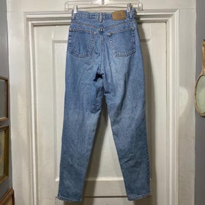 Puede incluir: Vaqueros de mezclilla azul claro con cintura alta y pernera recta. Los vaqueros tienen dos bolsillos traseros y un parche de cuero en la cintura. Los vaqueros están colgados de una percha de plástico transparente.