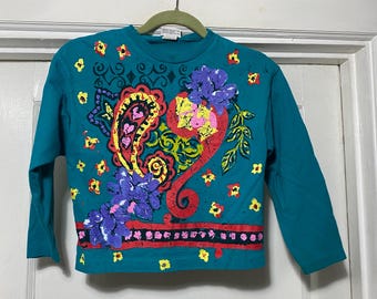 Puede incluir: Camiseta de manga larga de color turquesa con un colorido diseño de cachemira y flores. El diseño incluye detalles en rojo, amarillo, morado y verde. La camiseta está colgada de una percha verde.