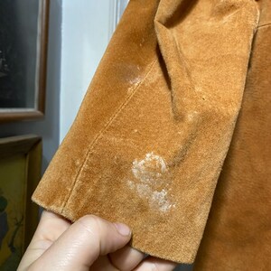 May include: Close-up of a tan suede jacket sleeve. The sleeve shows signs of wear with visible white discoloration. The jacket appears to be vintage, with a classic design. The texture of the suede is evident in the image.