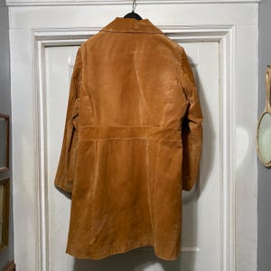 May include: A long, tan suede coat hangs on a black hanger against a white door. The coat has a classic design with a collar, buttons, and a tailored fit. The suede has a soft, textured appearance.