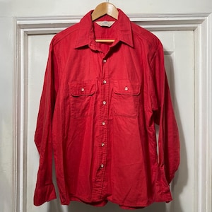 Puede incluir: Una camisa roja brillante de manga larga con dos bolsillos en el pecho y cuello. La camisa es abotonada y cuelga de una percha de madera. La tela parece ser un material de peso medio.