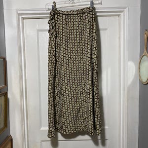 Puede incluir: Falda larga envolvente con un estampado floral repetido en negro, beige y verde claro. La falda está colgada de una percha contra una puerta blanca. La falda tiene un borde con volantes en la parte superior.