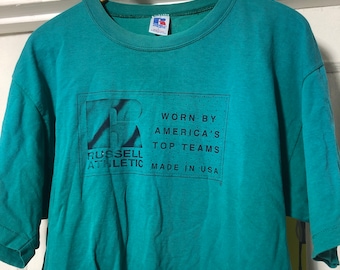 Könnte beinhalten: Türkisfarbenes Russell Athletic T-Shirt. Das Shirt hat eine schwarze Grafik mit dem Markenlogo und dem Text "Worn by America's Top Teams. Made in USA." Das T-Shirt hängt an einem Holzkleiderbügel.