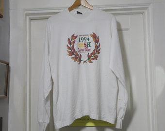 Könnte beinhalten: Weißes Langarmshirt mit einem Grafikdruck eines Lorbeerkranzes mit dem Text "1994 5K Spirit Run" darin.