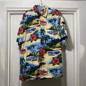May include: Short-sleeved button-down shirt with a tropical print. The design features red classic cars, palm trees, red hibiscus flowers, and a beach scene on a cream-colored background. The shirt is hanging on a hanger.