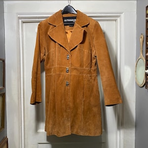 May include: A tan suede coat with a collared neckline and three decorative buttons. The coat has a fitted waist and long sleeves. The tag reads "OUTERWEAR". The coat is hanging on a black hanger against a white door.