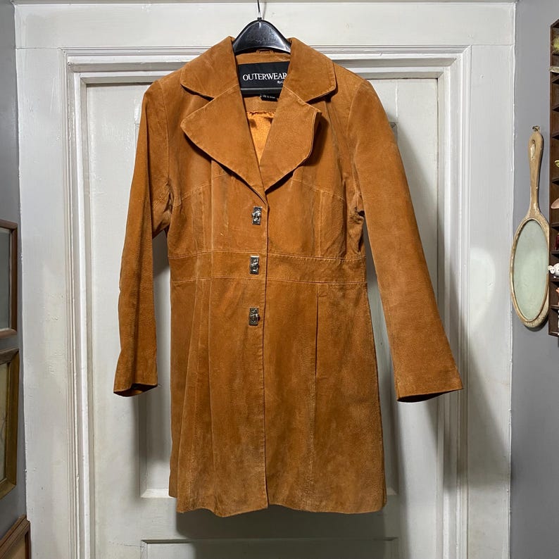 May include: A tan suede coat with a collared neckline and three decorative buttons. The coat has a fitted waist and long sleeves. The tag reads "OUTERWEAR". The coat is hanging on a black hanger against a white door.