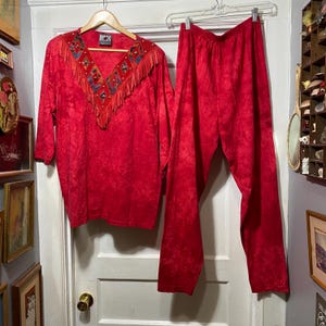 May include: A two-piece red outfit, consisting of a long-sleeved top and matching pants. The top features a V-neck with fringe and decorative elements. The outfit is hanging on a hanger against a white door.