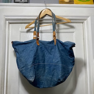 May include: A large, blue denim tote bag with brown leather accents on the straps. The bag is hanging on a wooden hanger. The denim has a worn appearance.