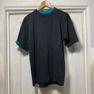 May include: Black short-sleeved t-shirt with a teal-colored crew neck and sleeve trim. The shirt is hanging on a wooden hanger against a white door. The shirt has a layered design.