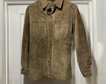 Può includere: Una camicia marrone in camoscio con due tasche frontali e un colletto. La camicia a maniche lunghe è appesa a una gruccia di legno. La camicia ha uno stile classico e casual.