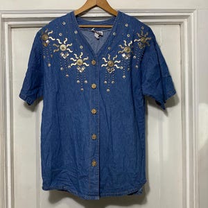 May include: A blue denim shirt with short sleeves and a button-up front. The shirt features a gold and silver sun design on the front.